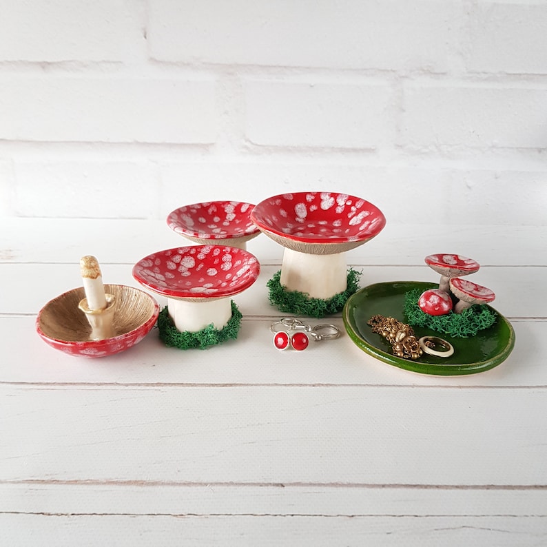 May include: A selection of ceramic mushroom-shaped ring holders and trays. The mushroom caps are red with white spots, and the stems are white. One ring holder is gold-coloured. A green tray holds jewellery and miniature mushrooms.
