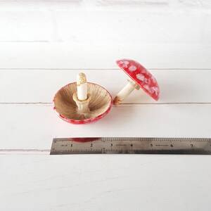 May include: Two ceramic mushroom ornaments. One is inverted, displaying the underside of the cap. The other is upright. Both have red caps with white spots and white stems. A ruler is in the foreground, showing measurements in inches.
