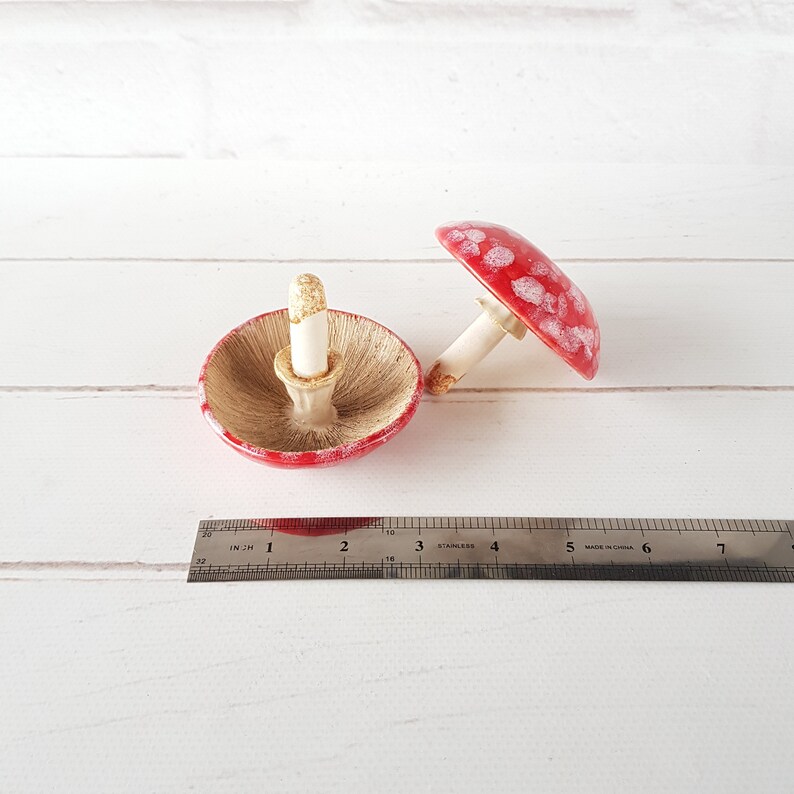 May include: Two ceramic mushroom ornaments. One is inverted, displaying the underside of the cap. The other is upright. Both have red caps with white spots and white stems. A ruler is in the foreground, showing measurements in inches.