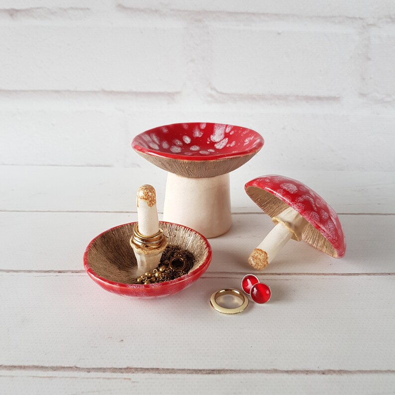 May include: Ceramic mushroom-shaped ring holders and trinket dishes. The red and white spotted mushroom caps sit atop beige stems. One ring holder displays gold rings. A gold ring and red stud earrings are also visible.