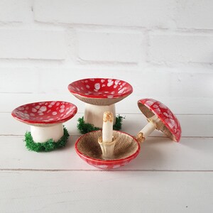 May include: Four ceramic mushroom ornaments. The mushroom caps are red with white spots, and the stems are white. One mushroom is upside down, showing the underside of the cap. The mushrooms are on a white surface.