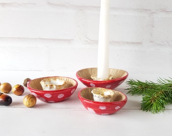 May include: Three red and white ceramic candle holders shaped like mushrooms. The candle holders have a white interior and a white dot pattern on the red exterior. A white candle is in the center holder.