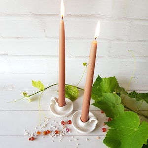 May include: Two white ceramic candle holders with brown candles burning. The candle holders are on a white surface with green leaves and small red and white stones.