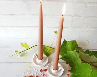 May include: Two white ceramic candle holders with brown candles burning. The candle holders are on a white surface with green leaves and small red and white stones.