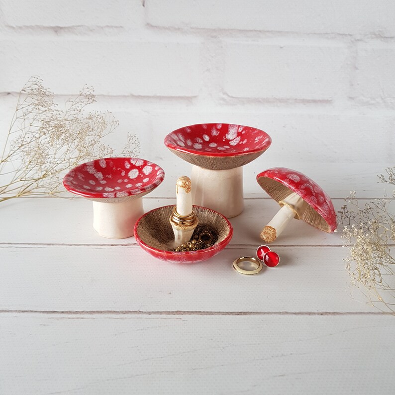 May include: A set of ceramic mushroom-shaped ring holders. The mushroom caps are red with white spots, and the stems are white. One holder contains rings and a lipstick. The background is a white brick wall and a white wooden surface.