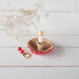 May include: A ceramic mushroom-shaped ring holder with a red cap and a beige underside. The stem is white with gold accents. Two rings and a pair of red stud earrings are displayed on a white wooden surface.
