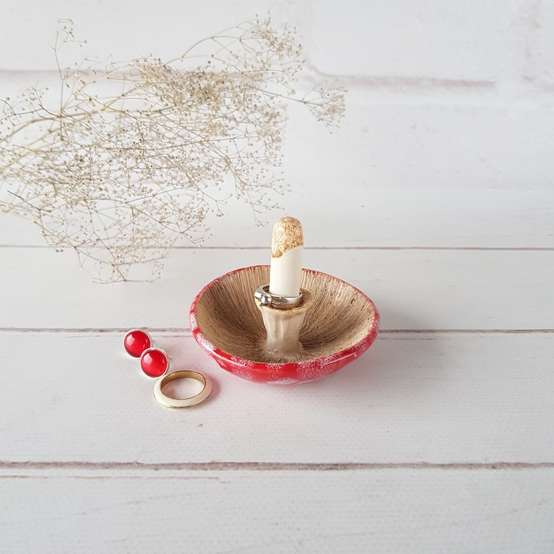 May include: A ceramic mushroom-shaped ring holder with a red cap and a beige underside. The stem is white with gold accents. Two rings and a pair of red stud earrings are displayed on a white wooden surface.