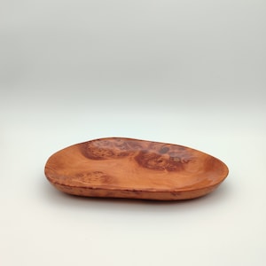 May include: A wooden serving tray with a natural reddish-brown colour. The oval tray has distinctive wood grain patterns and a smooth, polished finish. This decorative tray is suitable for serving snacks or displaying small items. The tray is approximately 30 cm long.