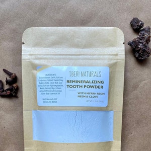 May include: A tan paper pouch of Iberi Naturals Remineralizing Tooth Powder with a blue powder visible through a window. The label lists ingredients and the text "WITH MYRRH RESIN, NEEM & CLOVE." Cloves and other natural ingredients are scattered around the pouch.