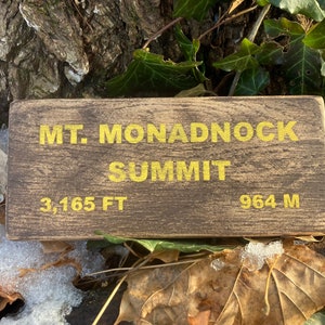 Puede incluir: Un letrero de madera con el texto "MT. MONADNOCK SUMMIT" en letras amarillas. El letrero también incluye la elevación en pies y metros: "3,165 FT" y "964 M".
