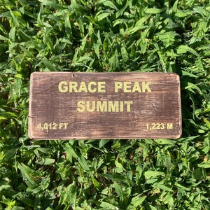Puede incluir: Un letrero rectangular de madera marrón con un acabado desgastado que presenta el texto dorado GRACE PEAK SUMMIT, 4,012 FT y 1,223 M. El letrero tiene una orientación horizontal con textura de veta de madera visible y bordes oscuros.