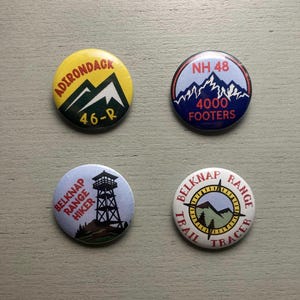May include: Four circular pinback buttons featuring hiking themes. One is yellow with a mountain and text reading ADIRONDACK 46-R. One is blue with a mountain and text NH 48 4000 FOOTERS. One is light blue with a fire tower and BELKNAP RANGE HIKER text. One is white with a compass and BELKNAP RANGE TRAIL RACER text.