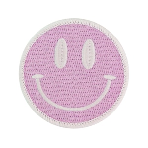 Peut inclure: Un écusson rond rose avec un visage souriant et une bordure blanche. Le visage souriant a des yeux ovales blancs et un sourire incurvé. L'écusson est fait d'un tissu texturé.