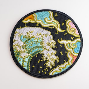 May include: A circular embroidered patch featuring a stylised ocean wave in white, light blue, and green against a black background. The design includes decorative clouds in shades of pink, yellow, and light blue with gold accents.