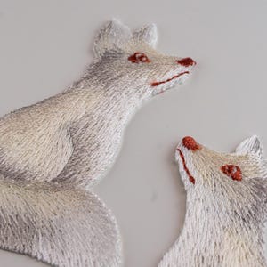 Könnte beinhalten: Zwei gestickte Aufnäher mit Fuchsmotiven in Weiß-, Creme- und Grautönen. Der größere Fuchs sitzt und blickt nach rechts, während der kleinere Fuchskopf darunter nach oben schaut. Beide haben Details aus rotem Garn an Augen, Nase und Mund.