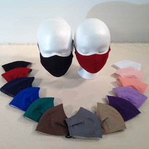 May include: A variety of colors of face masks are displayed on a white surface. The masks are made of fabric and have a variety of colors, including black, red, blue, green, brown, gray, pink, and purple. The masks are arranged in a circle around two mannequins wearing masks.