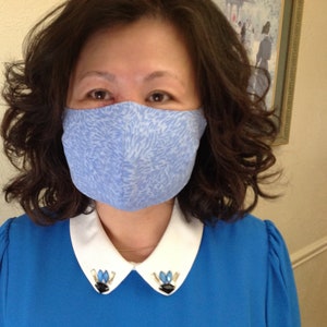 May include: A woman wearing a blue dress with a white collar and blue and white patterned face mask.