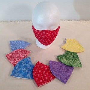 May include: A set of eight fabric face masks in various colors and patterns, including red, blue, green, purple, and yellow. The masks are arranged in a circle around a white mannequin head wearing a red mask with a white floral pattern.