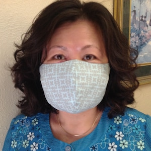 May include: A light blue face mask with a repeating pattern of gray "XOXO" symbols. The mask is worn by a person with dark hair and a blue top with floral embroidery. A gold necklace is visible.