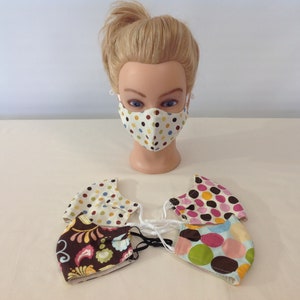 May include: Four fabric face masks with different patterns. One mask has a white background with brown, yellow, and blue polka dots. Another mask has a pink background with brown, yellow, and orange polka dots. A third mask has a brown background with a floral pattern in shades of pink, yellow, and green. The fourth mask has a light blue background with brown, pink, and green polka dots.