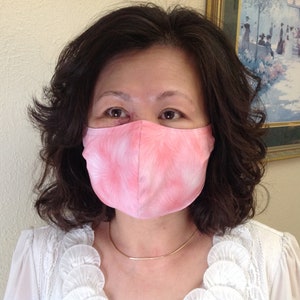May include: A pink face mask with a white, feathery pattern. The mask is worn by a person with dark hair and a gold necklace. The person is wearing a white blouse. The background includes a painting.