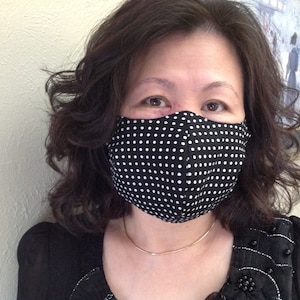 May include: A woman wearing a black and white polka dot face mask. The mask covers her nose and mouth. She is wearing a black top with silver embellishments.