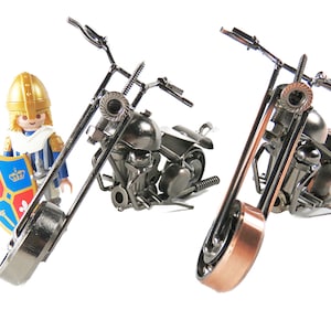 May include: Two metal motorcycle sculptures with detailed engine and frame components. One has a copper-colored front fork. A Playmobil knight figure stands nearby, holding a shield with a crest.
