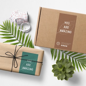Op de afbeelding: Twee bruine kartonnen dozen met een groene en bruine sticker waarop "You are amazing" staat met een bladerenontwerp. De dozen staan op een wit oppervlak met groene bladeren en een kleine vetplant.