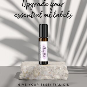 May include: A close-up of a 10 ml roller bottle with a white label that says "Rejuvenating Blend" in purple script. The bottle is sitting on a light gray stone surface. The text "Upgrade your essential oil labels" is at the top of the image. The text "Give your essential oil a professional makeover. Fit 10 ml roller bottles and more" is at the bottom of the image.