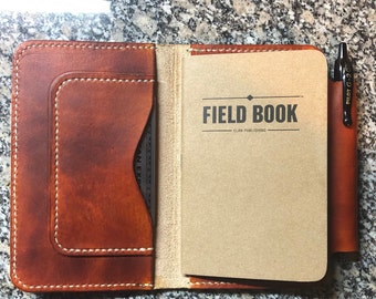 May include: A brown leather notebook cover with a pen holder and a pocket for a card. The notebook cover is open to reveal a blank, brown notebook with the text "FIELD BOOK - ELAN PUBLISHING" printed on the front.