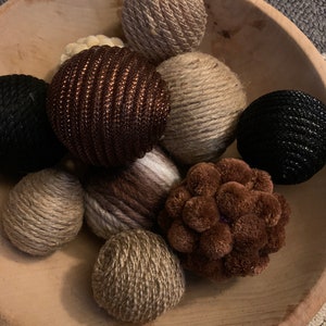 May include: A wooden bowl filled with various decorative balls. The balls are made of different materials, including twine, yarn, and fabric. The balls are in shades of brown, black, and white.
