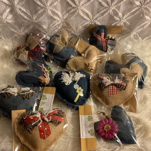 May include: A collection of handmade denim and burlap heart ornaments. The hearts are decorated with various embellishments, including flowers, bows, and ribbons. The ornaments are packaged in clear plastic bags with brown paper tags that read "Sheri's Country Crafts".