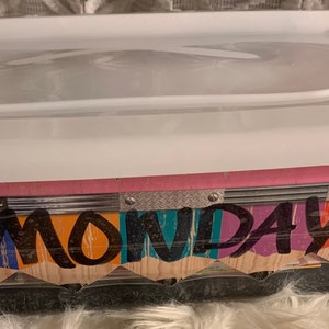 May include: A clear plastic storage container with a white lid. The container is decorated with colorful paper and the word "MONDAY" written in black marker.