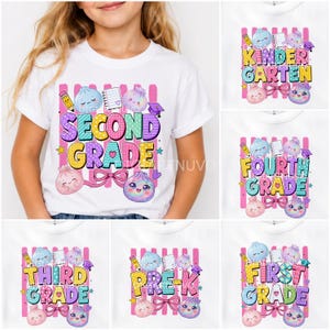 May include: A white short-sleeve t-shirt featuring a colourful graphic print with the text SECOND GRADE, surrounded by illustrations of smiling dumplings, a pencil, a notebook, and a graduation cap against a background of pink vertical stripes.