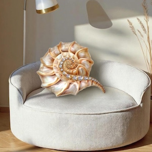 May include: A decorative pillow shaped like a nautilus shell with a pearlescent, light brown finish and a spiral of small, clear, sparkling rhinestones embedded along the center ridge of the shell, resting on a round, off-white textured chair.