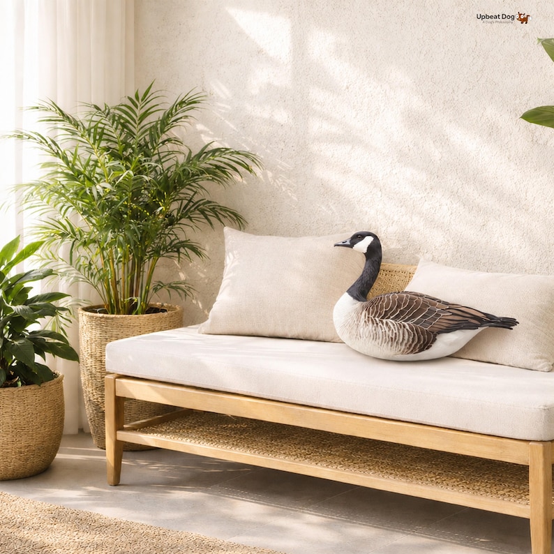 May include: A plush throw pillow shaped like a Canada goose with realistic brown, black, and white feather patterns. The pillow is resting on a light-colored bench cushion between two plain beige square pillows, with the text Upbeat Dog visible.
