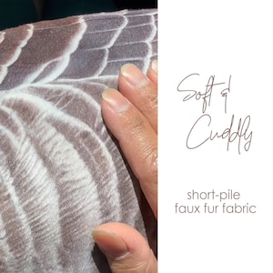 May include: A short-pile faux fur fabric featuring a repeating brown and white feather-like pattern. The text Soft & Cuddly and short-pile faux fur fabric is printed on the right side of the image in a dark brown script and sans-serif font.