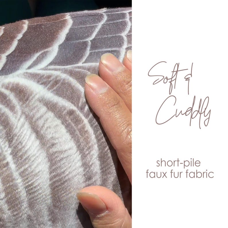 May include: A short-pile faux fur fabric featuring a repeating brown and white feather-like pattern. The text Soft & Cuddly and short-pile faux fur fabric is printed on the right side of the image in a dark brown script and sans-serif font.
