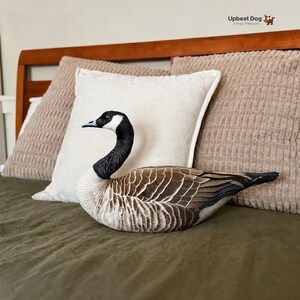 May include: A plush throw pillow shaped like a Canada goose with realistic brown, black, and white feather patterns. The goose has a black neck and head with a white cheek patch, a black beak, and a detailed wing design. The text Upbeat Dog A Dog's Philosophy is visible in the top right corner.