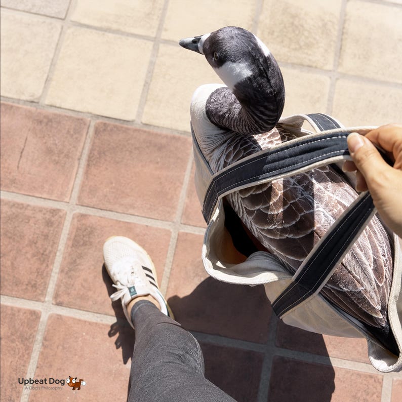 May include: A tote bag featuring a realistic, high-resolution print of a Canada goose. The bag is made of off-white canvas with dark grey fabric handles. The goose design covers the exterior, showing the bird's head, neck, and brown-feathered back.