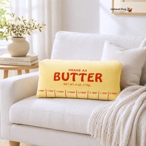 May include: A rectangular yellow throw pillow designed to look like a stick of butter. The front features red text reading GRADE AA BUTTER, NET WT. 4 OZ. (113g), and a measurement scale from 1 TBSP to 8 TBSP printed along the bottom edge.