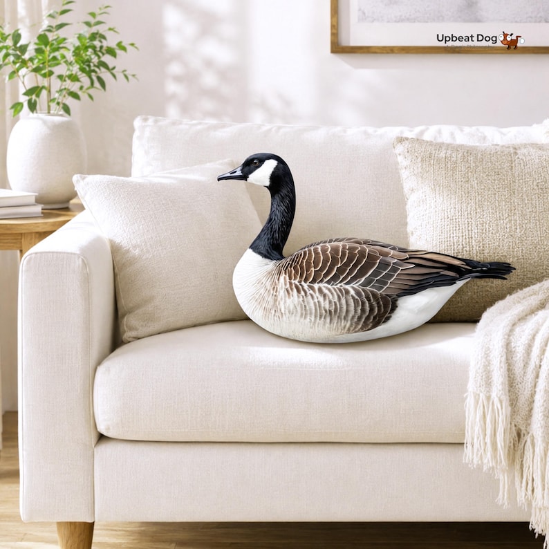 May include: A plush throw pillow shaped like a Canada goose with a black neck and head, white cheek patches, and brown, black, and white patterned wings. The pillow features realistic feather detailing and is positioned on a white sofa.