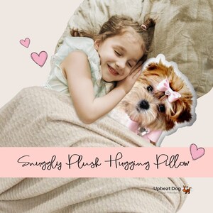 Op de afbeelding: Een knuffelkussen in de vorm van een kleine hond met een roze strik. Het kussen ligt op een bed met een jong meisje dat ernaast slaapt. De afbeelding heeft de tekst "Snuggly Plush Hugging Pillow" en "Upbeat Dog A Dog's Philosophy" erop.
