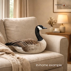 May include: A plush Canada goose toy featuring a black head and neck with a white cheek patch, a brown and tan patterned body, and a white underside. The goose is depicted in a resting position with its head held upright and alert.