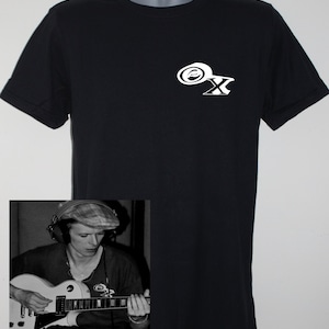 Op de afbeelding: Een zwart T-shirt met een witte afbeelding van een gestileerde letter X met een cirkel eromheen. Het T-shirt wordt gemodelleerd door een persoon die een zwart-wit gestreept shirt en een zwarte pet draagt. De persoon speelt een witte elektrische gitaar.