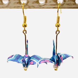 May include: A pair of origami crane earrings with blue, pink, and white paper. The cranes are hanging from gold hooks with pearl beads at the bottom.