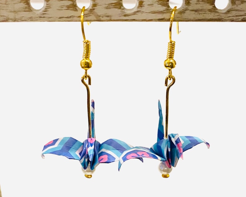May include: A pair of origami crane earrings with blue, pink, and white paper. The cranes are hanging from gold hooks with pearl beads at the bottom.