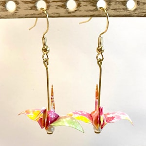 Può includere: Un paio di orecchini pendenti color oro con disegni di gru origami. Le gru sono realizzate in carta colorata con sfumature rosa, gialle e verdi. Ogni orecchino ha una piccola perla sul fondo.