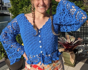 May include: A blue crocheted top with long sleeves and a button closure. The top has a scalloped hem and a delicate lace pattern.