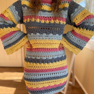May include: A colourful crochet dress with a striped pattern in shades of yellow, pink, blue, and grey. The dress has a loose fit and long sleeves with a bell shape.